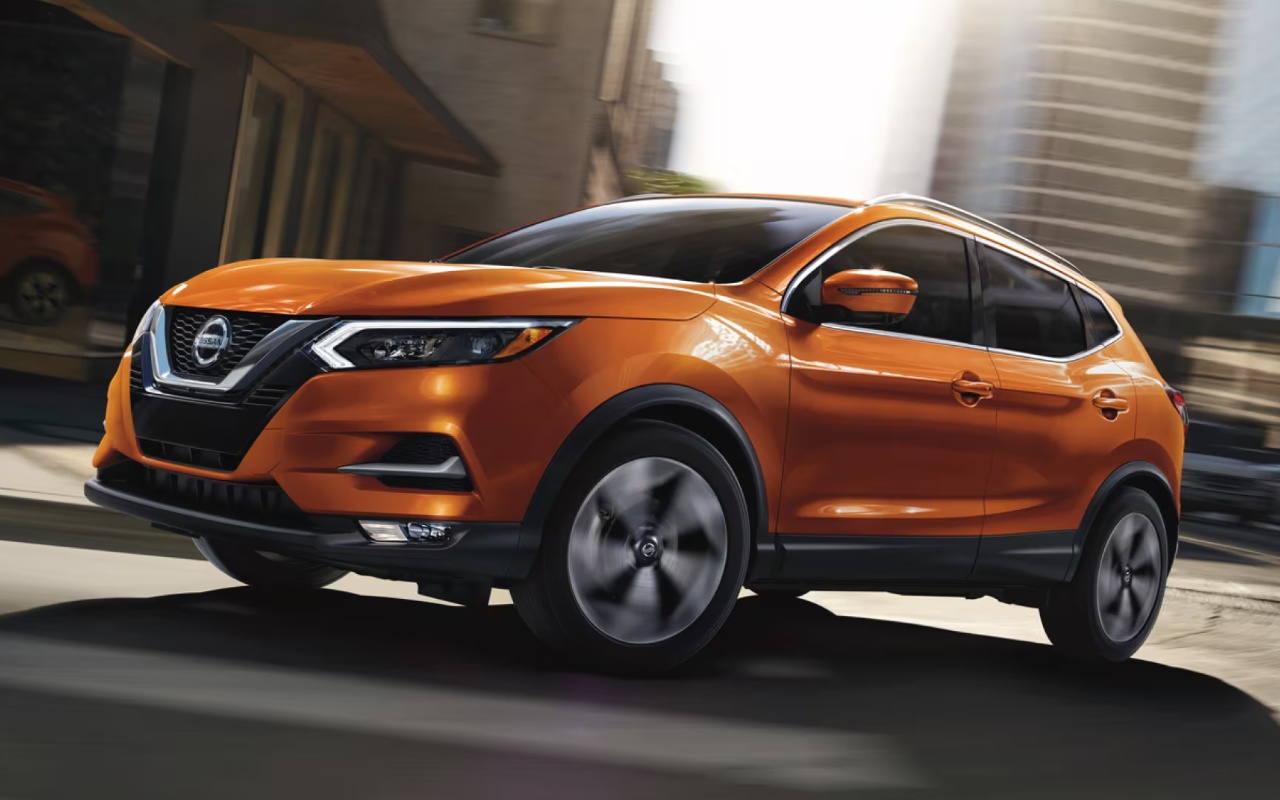 Nissan Qashqai orange de génération précédente en mouvement dans une rue de centre-ville ensoleillée.