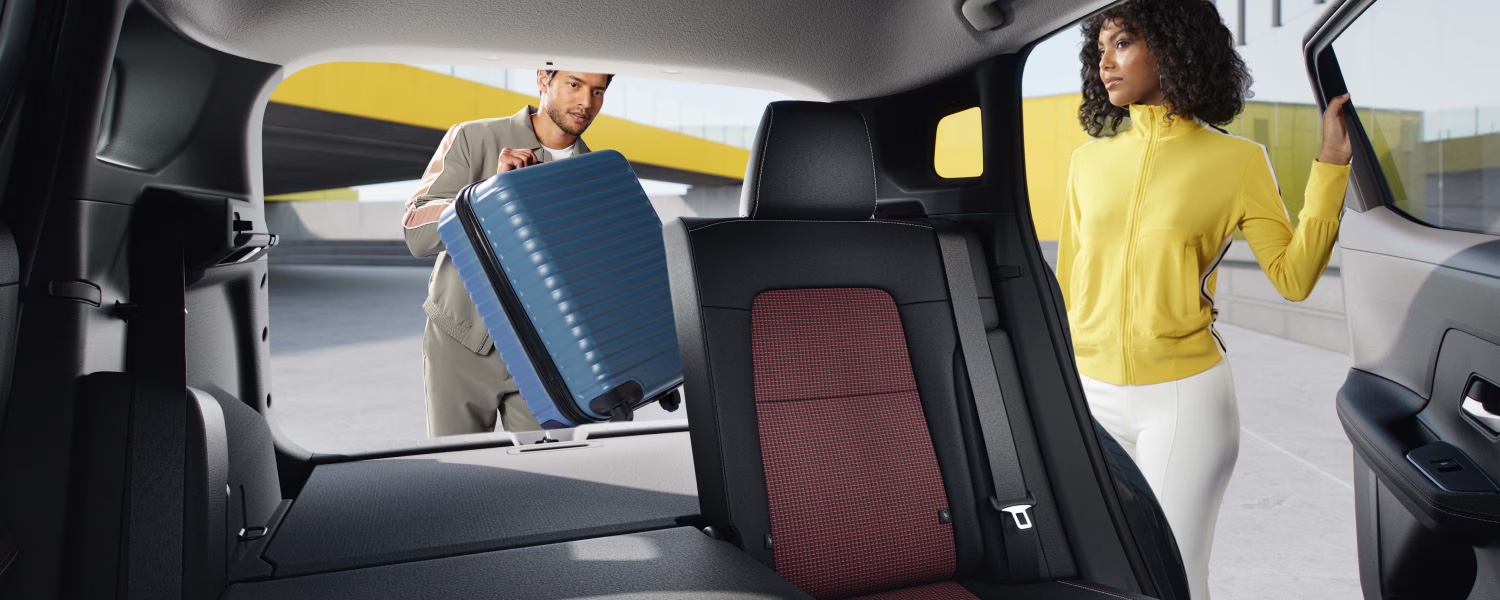 Vue intérieure du Nissan Kicks 2025 avec femme debout à l’arrière et homme rangeant une valise bleue dans le coffre.